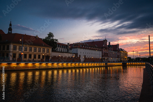 Scenic evening view of a historic European city embankment at sunset, with old buildings and streetlights reflecting in the river. Travel and tourism concept	
