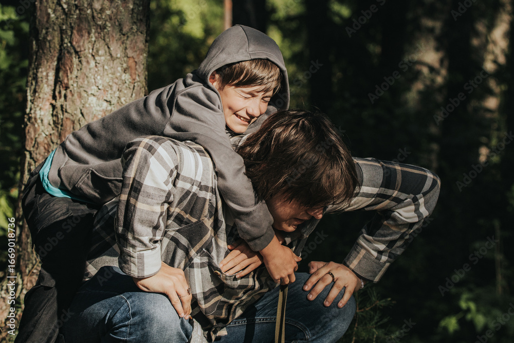 Fototapeta premium A boy in a hoodie hugs an older boy in a plaid shirt from behind in a forest, sharing a tender and playful moment in the sunlight.
