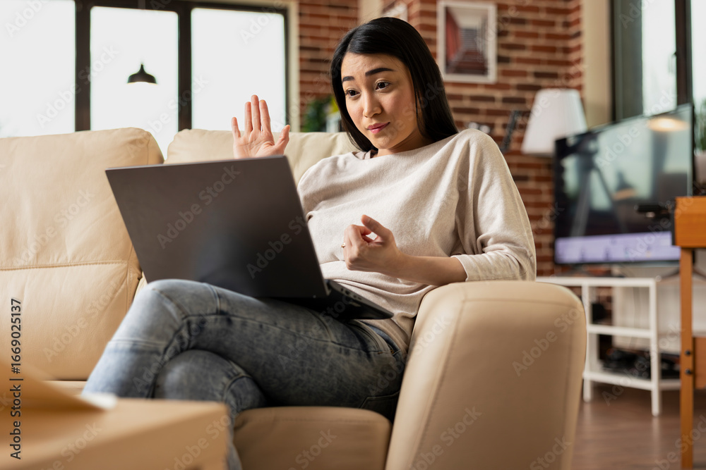 Fototapeta premium Smiling asian businesswoman waving hello at laptop during remote meeting, holding coffee cup in home office. Female virtual assistant greets coworkers through online video conference on digital device