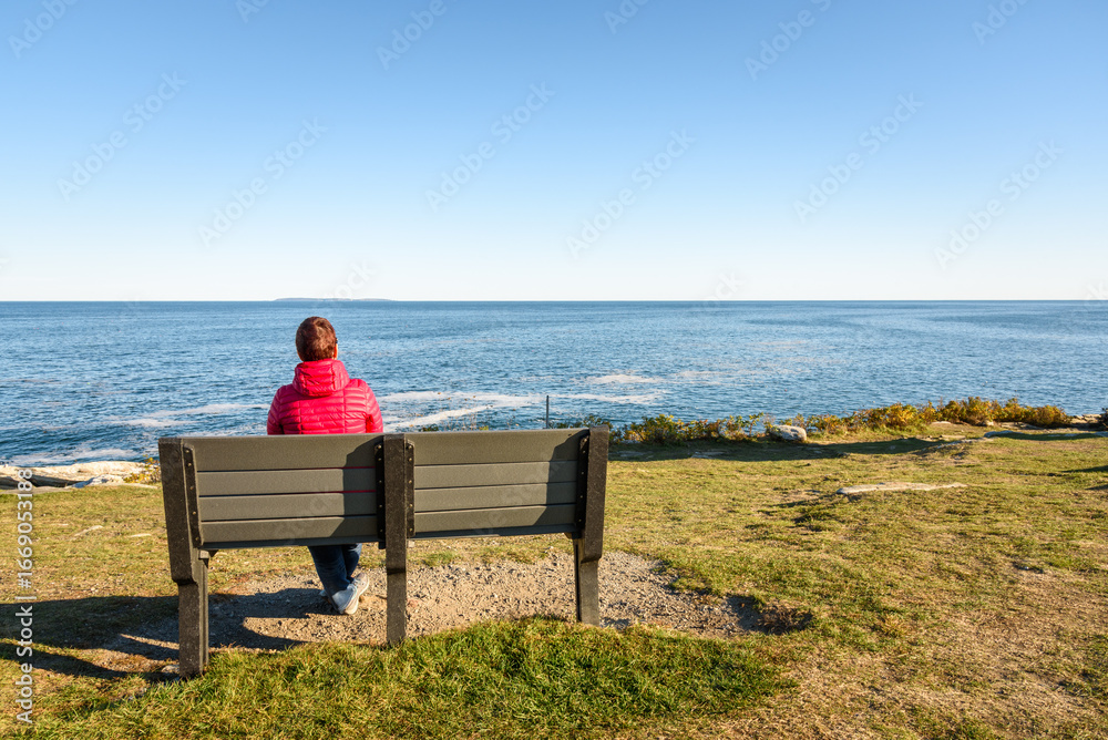 Fototapeta premium Woman enjoying a magnificent seascape while sitting on a bench on the top of a cliff on a clear autumn day. Pemaquid Point, ME, USA.