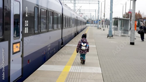 Small child in winter jacket with penguin backpack walking alone on empty train station platform next to passenger train. Family travel, public transportation and childhood independence journey. 