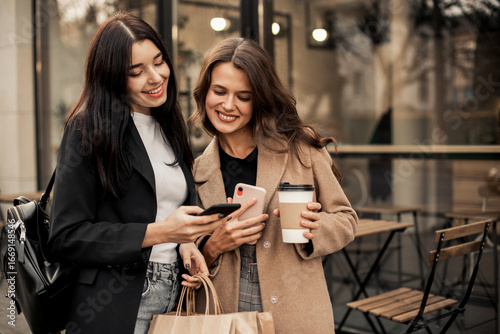 Two smiling women friends dressed in beige coat and jacket drinking coffee and using smartphone or phone, holding paper bags with purchases in hand after shopping walking along streets of autumn city.
