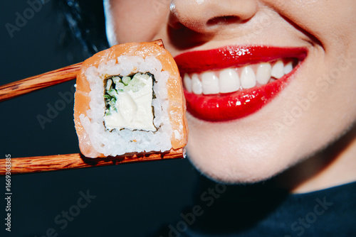 Young woman with red lips smiling while holding sushi with chopsticks, showcasing vibrant colors and a modern dining concept.