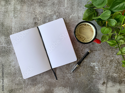Top view of stone top desk workspace with an open mock-up friendly blank notebook with space for text overlay and pen next to it, a hot cup of coffee and a green plant in view.
