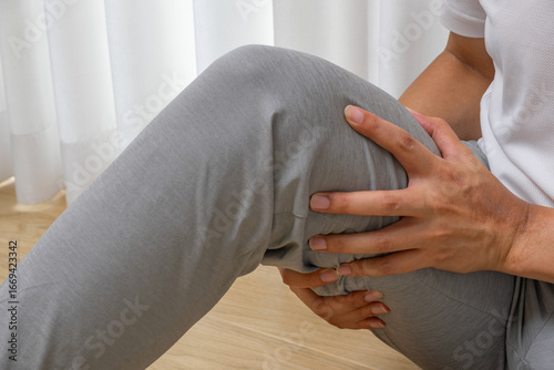 Asian man seated indoors, clutching hamstring in post-run pain, possible muscle strain or tightness. Morning exercise, sports injury, rehab, wellness, medical care.