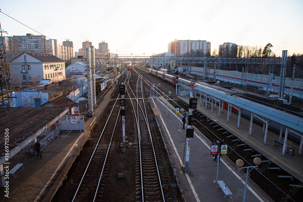 Fototapeta premium A railway station with multiple tracks and platforms. Buildings and urban structures are visible in the background. The scene is illuminated by sunset light.