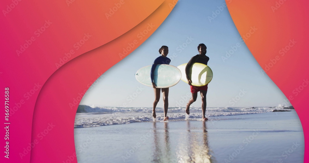 Fototapeta premium Walking couple wearing wetsuits carrying surfboards on wet beach, with waves reflecting on sand