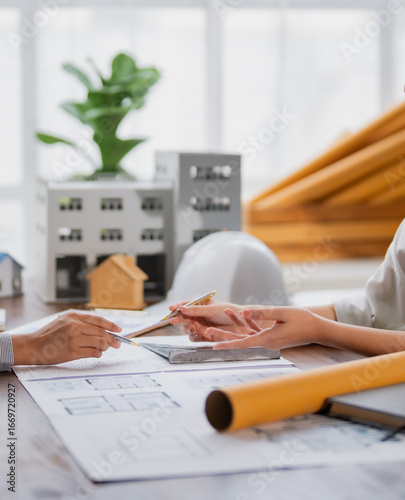 Two architects discussing new project details while examining blueprints and a scale model of a building, collaborating effectively in a modern office environment