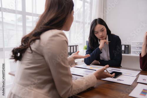 Asian businesswomen are discussing marketing strategy holding smartphone and analyzing financial statistics displayed on documents and graph chart during a meeting in a modern office
