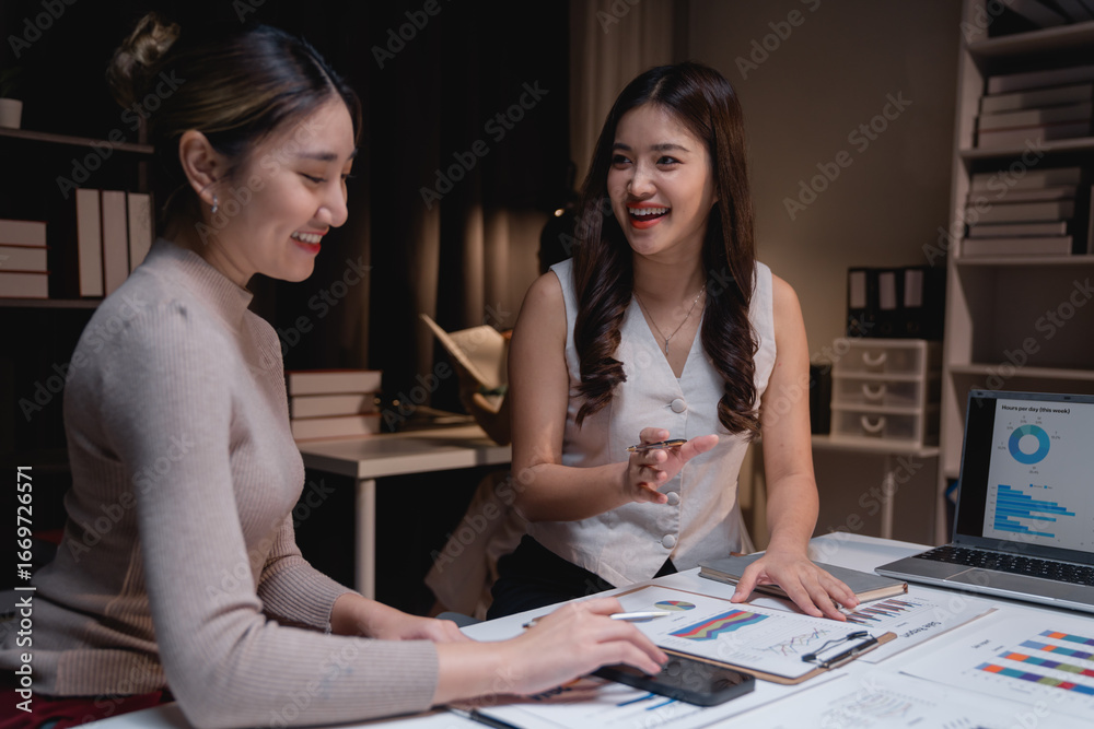 Fototapeta premium Two smiling Asian businesswomen are discussing a marketing strategy using charts, graphs, and a laptop, working late at night in the office to achieve their business goals