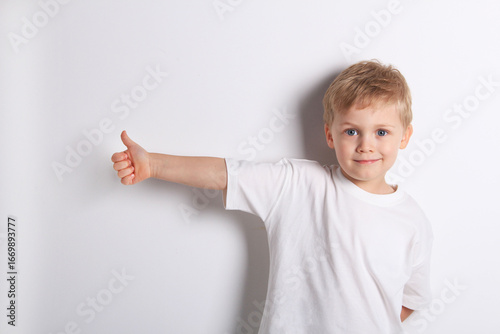 Cute little boy in white T-shirt gives thumbs up on white background 