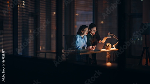 Two professionals cooperating project overtime working at laptop in lamp light. 