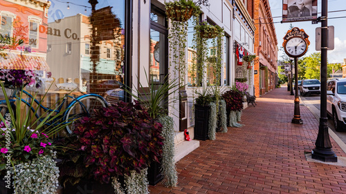 West Union, United States - 13 August 2025: View of a quaint street with brick buildings, flower-filled planters, and a vintage clock casting shadows on the red brick pavement.