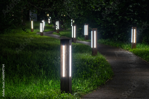 Fotografía Tranquil nighttime park path illuminated by series of modern, rectangular bollard lights, casting serene glow on surrounding green grass and dark foliage