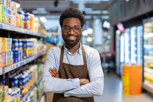 Konstfotografi Smiling employee in grocery store aisle wearing apron while standing confidently