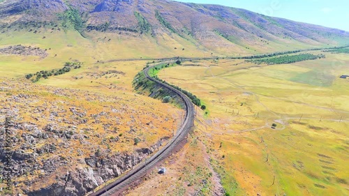 Railway curving through hilly plains at the foot of mountains. Aerial view under the blazing sun