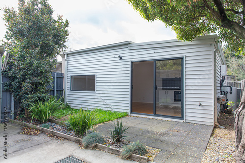 Exterior view of modern compact home with sliding glass door leading to patio, white siding walls and small landscaped garden area