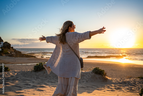 Beautiful mature woman standing with outstretched arms on sandy beach on sunset on spring day. Back view. Monte Clerigo beach on Algarve coast in Portugal	