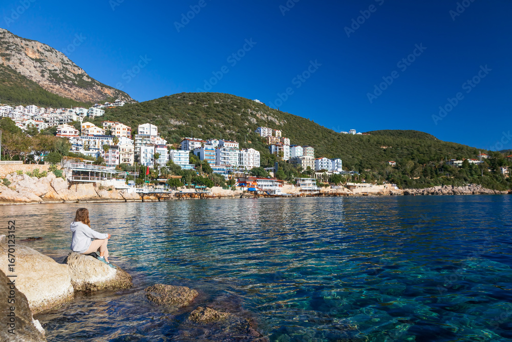 Obraz premium Woman looking at sea and historical town Kash placed on hills by coast of mediterranean sea in Antalya region in Turkey. Capturing summer vacation, popular place for family travel with kids.