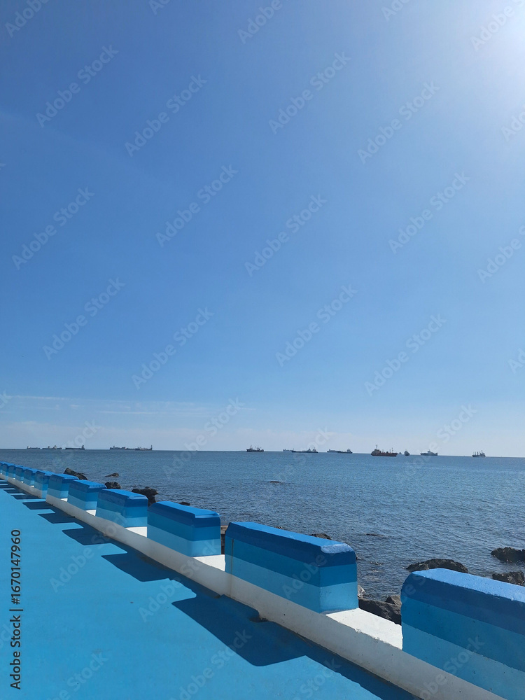 Obraz premium Scenic ocean view with blue painted seawall and clear sky, ships floating on horizon, bright sunny day by the coast, peaceful seaside landscape with vibrant colors and calm summer atmosphere