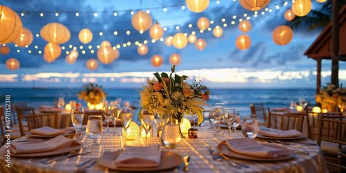 A beautifully arranged outdoor dining table with elegant decorations. Soft glowing lanterns hang above, creating a warm ambiance by the beach at dusk.
