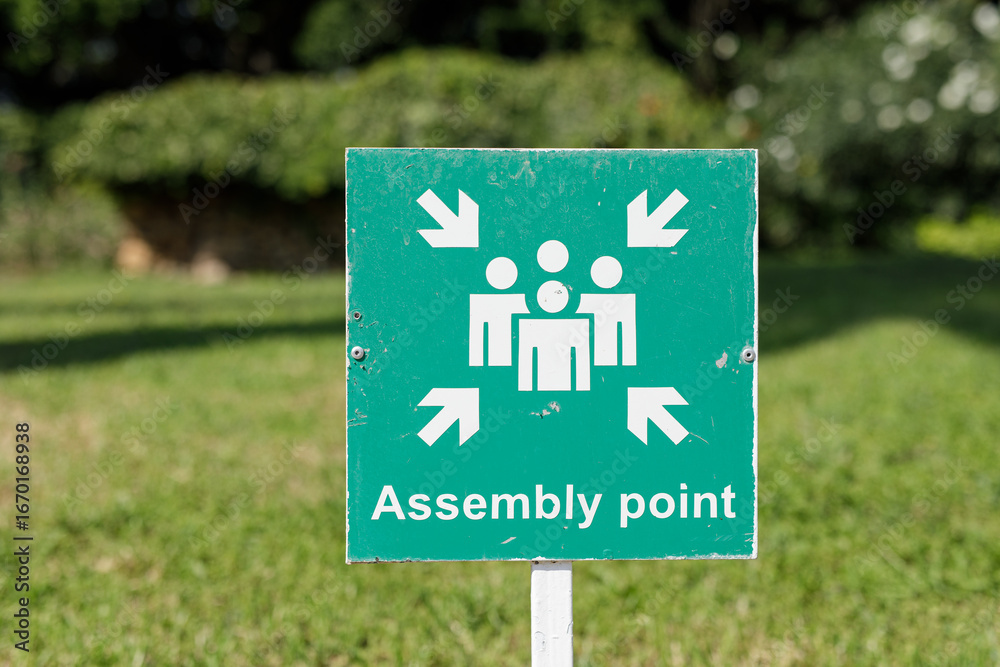 Φωτογραφία Stock photo of a green assembly point sign with arrows indicating direction, situated outdoors on a grassy lawn