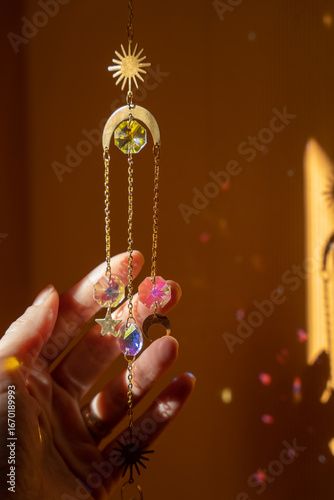 Glass suncatcher with metal golden chains, a woman hand touches it, shining in sunlight and reflecting the rainbow on yellow background