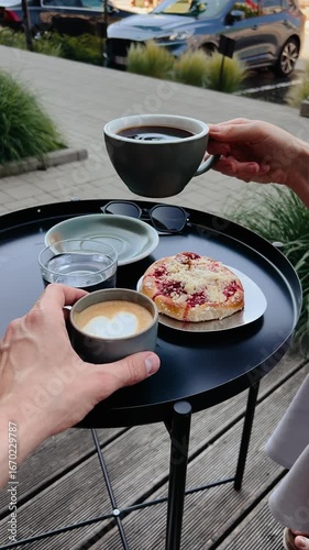 First-person view: two people are sharing small round table with two cups of coffee, glass of water, small season bun on small plate, outdoors in cafe. Two person sharing coffee, water, cake outside