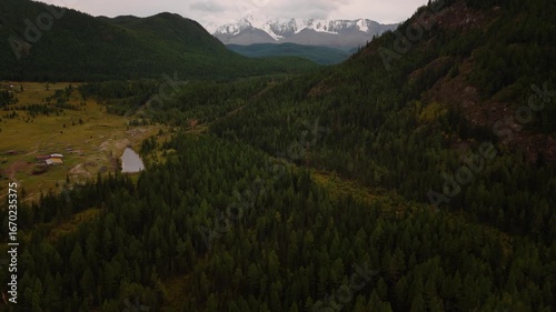 Scenic aerial view of mountain valley with forest, meadows, and ponds. Snow-covered peaks in distance under dramatic sky. Ideal for nature, adventure, and environment footage.
