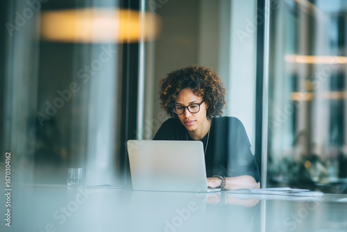 Focused woman working on laptop in modern office, wearing glasses with curly hair, showcasing concentration and professionalism.