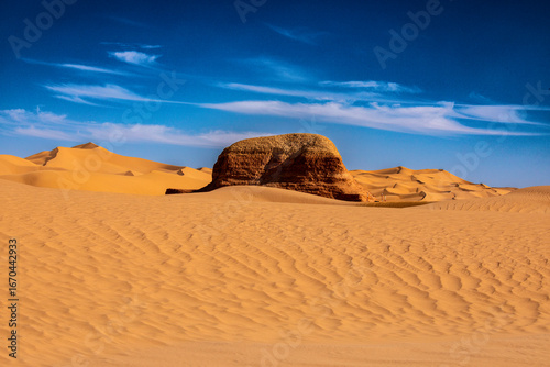 Fotografia A large, reddish-brown rock formation with a textured top sits prominently in the foreground of a sandy desert