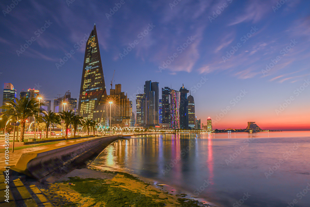 Obraz premium Doha city business downtown center panorama illuminated at sunrise, with modern skyscrapers and sea in the foreground, Qatar