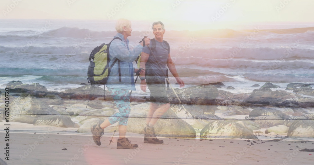 Obraz premium Using trekking poles couple hikers walking along beach at sunrise, with backpacks