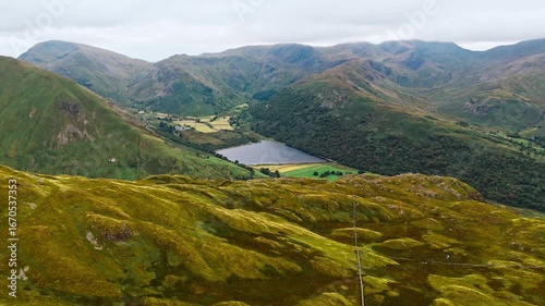 aerial drone shot high angle over mountain valleys with brothers water lake in centre of shot slowly panning right in Lake District England in summer with cloudy skies