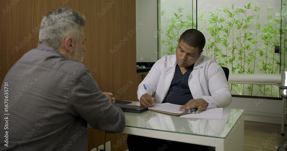 Obraz premium Male doctor writing medical notes during consultation with middle-aged patient seated at desk in clinic, both focused on conversation and paperwork