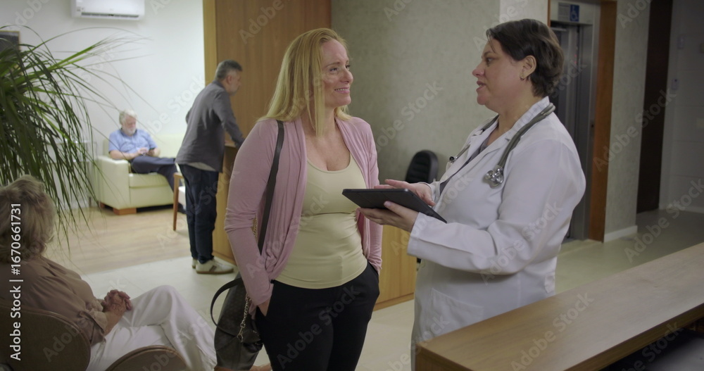 Obraz premium Female doctor speaking with woman patient near clinic reception, hand on shoulder while reviewing information on tablet in warm healthcare environment
