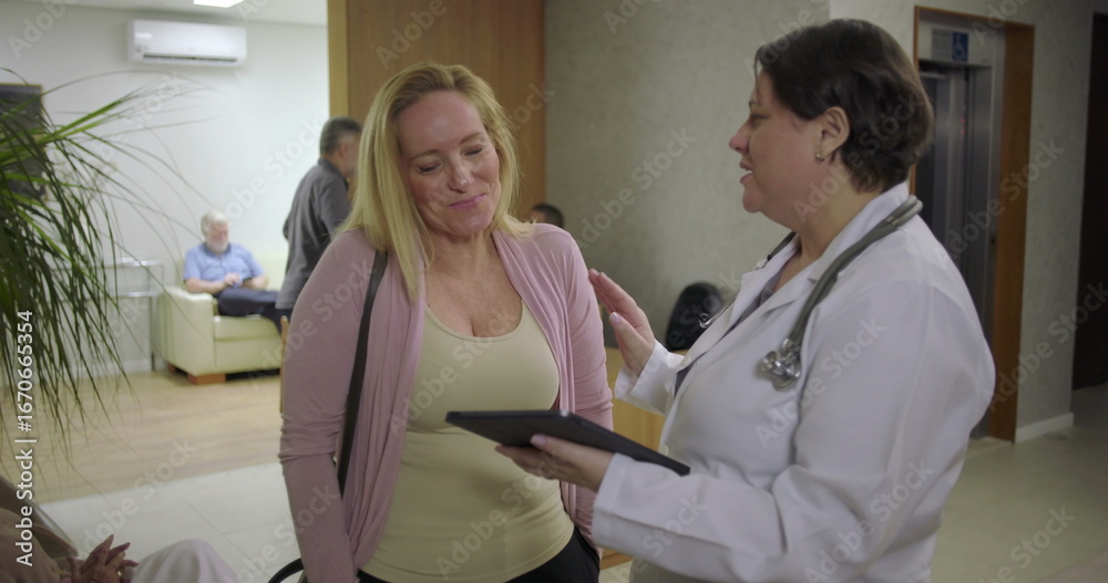 Fototapeta premium Female doctor reviewing medical details on tablet with woman in reception area of clinic, positive healthcare conversation near waiting area
