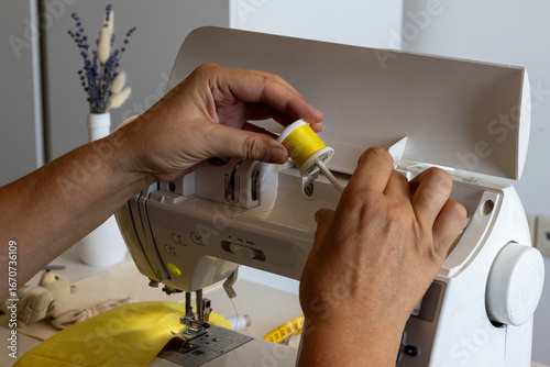 Wallpaper Mural Close-up of a person's hands threading a white sewing machine with a bright yellow spool of thread. Yellow fabric is visible under the needle, suggesting a creative project. Torontodigital.ca
