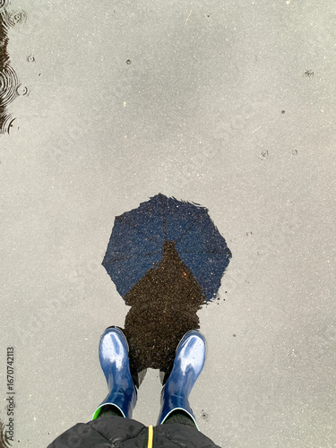 A reflection on a water puddle of a person holding an umbrella during a rainy day and a view of the rain boots for inclement weather concept with copy space. 