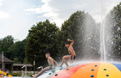  Two children sliding and splashing down colorful water dome attraction with fountain spray at outdoor aquatic center during cloudy summer day. Water park thrills, sibling fun, recreational activities