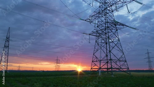 Power lines and pylons silhouetted against a vibrant sunset in a rural field, symbolizing sustainable energy infrastructure and harmony with nature, captured in a long shot.