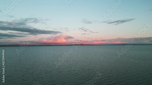 Rising high above to reveal wind turbines along the edge of the shore with a stunning sunset and lake in the foreground.