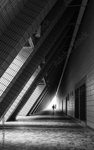 Photography A lone figure walks down a long, angled hallway under dramatic shadows and light, surrounded by textured walls and repeating geometric patterns in this black and white architectural perspective
