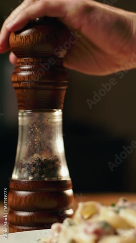 Hand grinding pepper with a wooden and glass pepper grinder over a food dish on a wooden table
