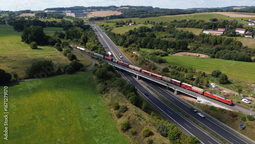 Drone view of freight train crossing bridge over busy highway in rural countryside with fields and hills