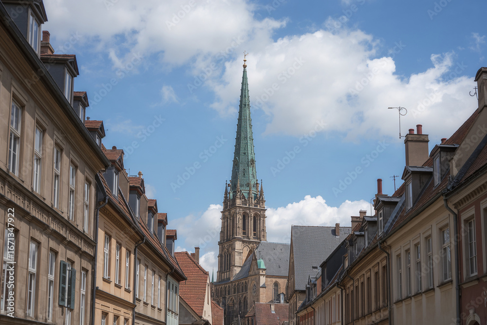 Obraz premium Tall green spire church between old European buildings under a cloudy sky architecture
