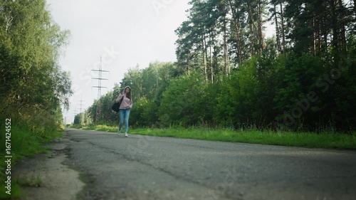 Wallpaper Mural Young lady in pink hoodie walking along quiet forest road with one hand in pocket and black bag on shoulder, surrounded by lush greenery, trees, and distant power poles under soft daylight Torontodigital.ca