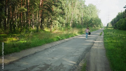 Wallpaper Mural Remote female worker in pink hoodie and jeans walks alone on cracked rural road through green forest, calmly holding black bag in hand, surrounded by nature and power lines Torontodigital.ca