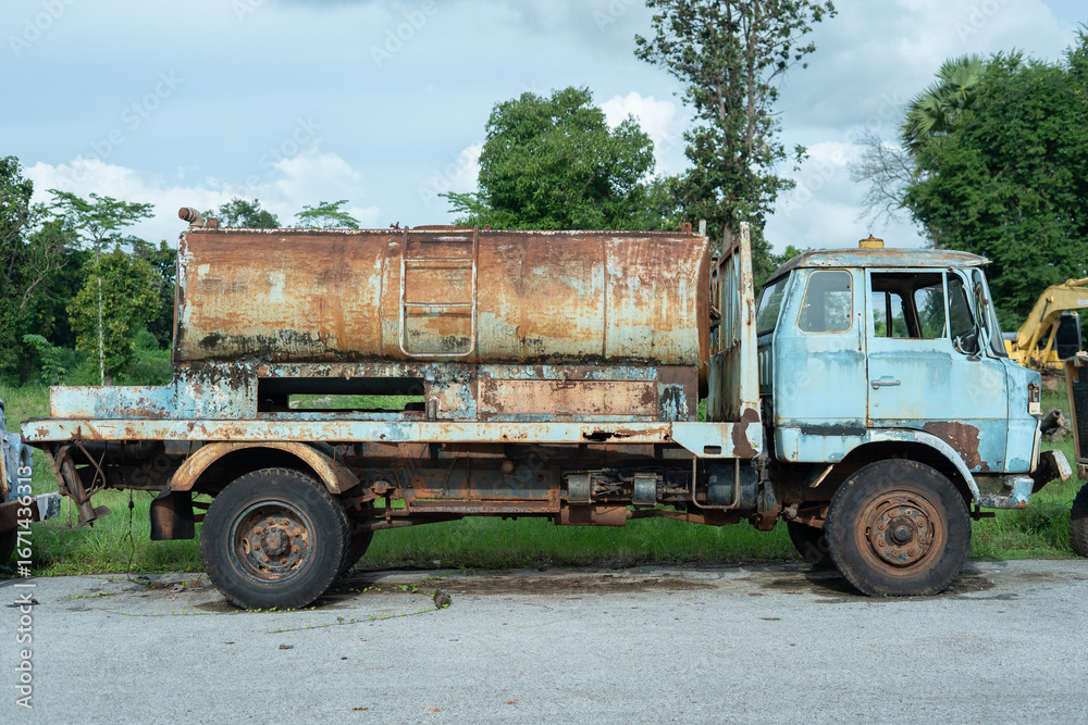 Fototapeta premium Rusty Truck: A weathered, rusty truck sits parked, its paint peeling and metallic surfaces worn by time, against a backdrop of greenery and a cloudy sky.