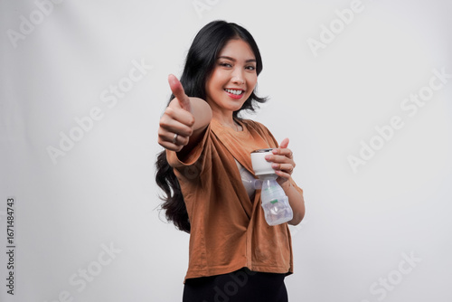 Pretty Asian mother pumping breastmilk with breast pump machine while doing thumb up gesture.